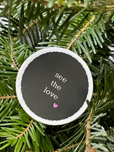 Load image into Gallery viewer, black, 1.5&#39;, round sticker with white border and white message, see the love, with a small deep pink heart at the bottom right of the letters 
