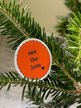 Load image into Gallery viewer, orange, 1.5&#39;, round sticker with white border and black message, see the love, with a small black heart at the bottom right of the letters 
